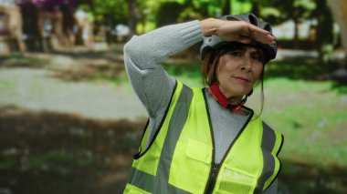 Miğfer ve yansıtıcı yelek giyen kıdemli İspanyol kadın parktaki mesafeye bakıyor. Etrafını çevreleyen ağaçlar ve yeşillikler, dış güvenlik ve yaşlı aktiviteleri somutlaştırıyor..