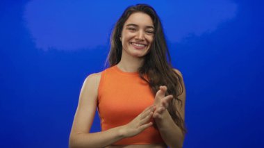 Mavi stüdyoda el çırpan, turuncu bluz giyen, geniş bir gülümsemeyle gülümseyen genç İspanyol kadın..