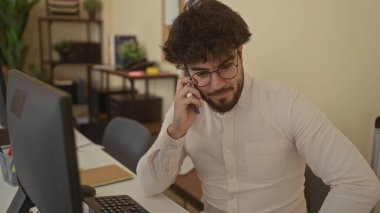 Ofiste sakallı genç adam telefonla konuşuyor. Masalar, bilgisayarlar ve bitkilerle çevrili. Bu da modern ve profesyonel bir çalışma alanı olduğunu gösteriyor..