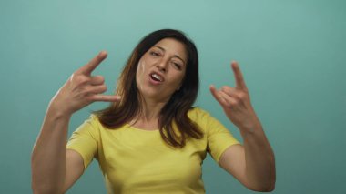Kaya boynuzları yapan kadın, elleri havada, sarı tişörtlü, gök mavisi stüdyo ortamında, rock-fun metal enerjili..