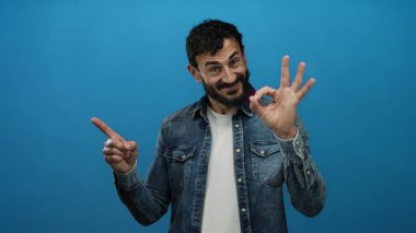 Sakallı, kot tişörtlü İspanyol bir adam. Tamam işareti yapıyor. Parlak mavi arka plana gülümsüyor..