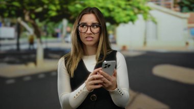 Gözlüklü bir kadın güneşli bir sokakta akıllı telefon kullanıyor. Arka planda ağaçlar ve binalar olan sıradan bir açık hava ortamında düşünceli görünüyor..