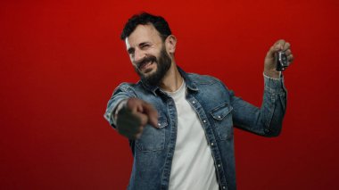 Kot ceketli İspanyol adam heyecan verici bir şekilde elinde kırmızı arka planlı araba anahtarı tutuyor. Promosyon amaçlı dinamik ve canlı bir atmosfer yaratıyor..