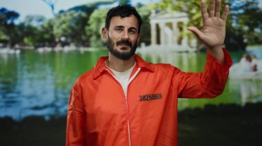 Turuncu tulumlu, sakallı, yeşil bir parkta ağaçlarla çevrili bir yerde elini kaldıran İspanyol bir adam..