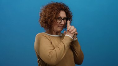 Latin bir kadın, parlak mavi bir duvara karşı silah hareketi yapıyor. Kendine güveni ve mizahı ifade ediyor. Gözlük takıyor ve kahverengi bir süveter giyiyor..