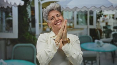 Kısa gri saçlı kadın, restoran terasında yumruğunu sıkar ve gülümseyerek yanaklarına dokunur; zaferi kutlar..