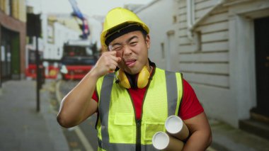 İnşaat malzemesi giymiş genç bir adam şehir sokaklarında alay ediyor, elinde planlar, sıkı bir şapka ve güvenlik yeleği var, şehir mimari temalarını çağrıştırıyor..