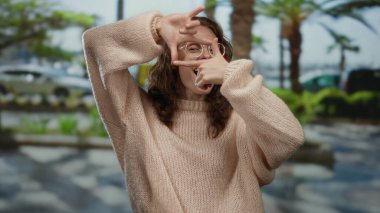Parkta bir kadın fotoğraf jesti yapıyor, gözlük ve süveter takıyor, açık havada ağaçlarla ve palmiyelerle gülümsüyor, doğal olarak yakalanan sıradan ve neşeli bir anı işaret ediyor..