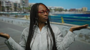 Gözlüklü kadın plaj gezintisinde huzur içinde poz veriyor. Arka planda deniz kenarı var. Kıvırcık saçlar, sakin bir ifade ve sessiz tonlar sergiliyor..