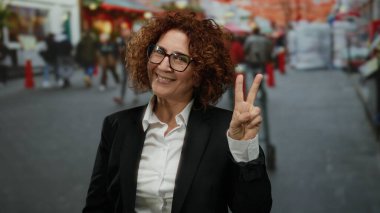 Kıvırcık saçlı, gözlüklü, gülümseyen ve barış simgesi olan bir kadın. Yoğun bir şehir caddesinde, arka planda yürüyen insanlarla birlikte..