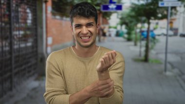 Kırık bilekli bir adam kalabalık bir sokakta kolunu tutarken dışarıdaki Latin kökenli genç adam bilek sakatlığından muzdarip..