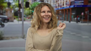 Dalgalı saçlı bir kadın, kalabalık bir caddede sıcak bir gülümsemeyle poz veriyor. Sarışın bir eşarp ve şehir ortamında genç bir stil takıyor. Açık havada mutlu görünüyor..