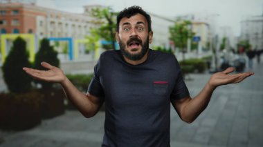 Arka planda bulanık binalar olan bir sokakta belirsizlik gösteren sakallı İspanyol bir adam..