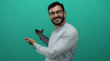 Sakallı, neşeli yeşil bir arka plana gülümseyen, el kol hareketi yapan, gözlüklü, açık mavi gömlekli, neşe saçan, parlak bir ortamda hoş geldin diyen bir adam..