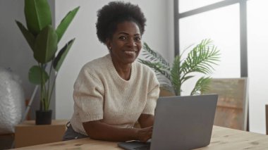 Modern oturma odasında dizüstü bilgisayar kullanırken gülümseyen bir kadın. Etrafı bitkiler ve kutular ile çevrili. Rahatlamış ve rahat bir atmosferi olan yeni bir ev ortamı öneriyor..