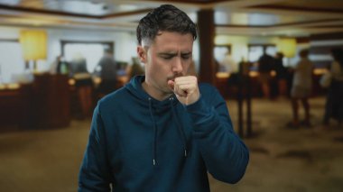 Mavi kapüşonlu genç adam arka planda yürüyen insanlarla dolu yoğun bir otel lobisinde öksürüyor..