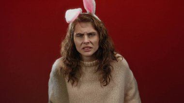 Kahverengi saçlı, tavşan kulağı takan, izole edilmiş kırmızı arka planda rahat süveter içinde kafa karışıklığı gösteren bir kadın.