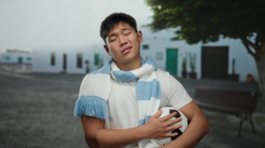 Beyaz gömlekli genç adam futbol topunu dışarıda tutarken aynı zamanda şehir caddesinde ağaca yaslanıyor ve gün boyunca sakin bir ifadeyle eşarp takıyor.