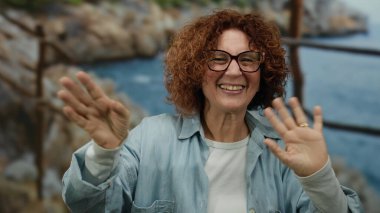 Okyanus manzaralı arka planda, kumral saçlarıyla sahilde kayaların arasında neşeli bir şekilde gülümseyen bir kadın..