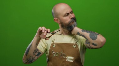 Dövmeli sakallı adam elinde makasla yeşil arka plana önlük takıyor. Yorgunluğunu ve düşüncelerini dile getiriyor..