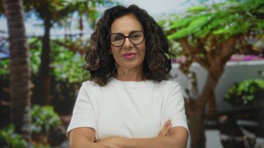 Beyaz tişörtlü İspanyol orta yaşlı kadın, yeşil parkta palmiye ağaçlarının altında çıplak kollarını kavuşturuyor; reddediliyor..