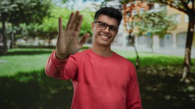 Açık hava parkında gülümseyen gözlüklü genç adam güneşli bir günde kırmızı kazak giyiyor. Etrafı yemyeşil ve şehir planlarıyla çevrili. Keyifli bir ortam izlenimi veriyor.