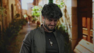 Sakallı genç adam canlı bir sokak manzarasında başını tutarak stresli ve düşünceli bir şekilde etrafı bitkilerle ve kır evleriyle çevrili duruyor..