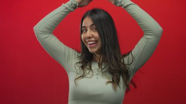 Açık yeşil bluzlu İspanyol kadın kırmızı stüdyo duvarına karşı elleri havada çatı jesti yaparken gülümsüyor..