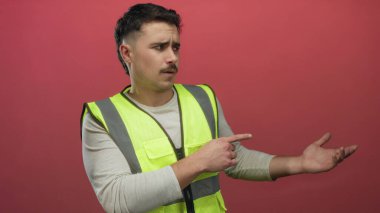 Yansıtıcı yelek giymiş genç İspanyol bir adam, izole edilmiş kırmızı arka plan duvarına işaret ediyor, odaklanmış bir ifadeyle yönü gösteriyor..