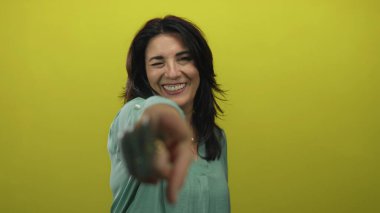 Gülümseyen orta yaşlı İspanyol kadın, sarı stüdyo seti içinde parmaklarını kameraya doğrultarak deniz mavisi bluz giyiyor..