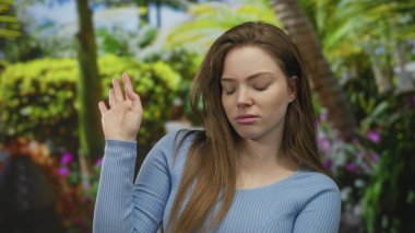 Genç Kafkasyalı kadın, tropik yeşilliklerin ortasında yeşil parkta el sallıyor ve yüzünü buruşturuyor; iğrenme.