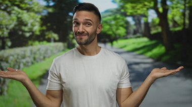 Parlak güneş ışığı altında yemyeşil bir parkta neşeli hareketler yapan genç adam, arka planda ağaçlar ve patikalarla tasasız ve neşeli bir hava yayıyor..