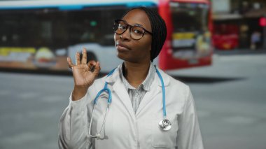Şehir merkezindeki kadın doktor, kameraya güvenle gülümseyen beyaz önlüklü ve gözlüklü, steteskoplu iyi bir jest yapıyor.