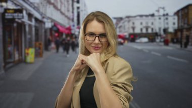 Gözlüklü, kendine güvenen sarışın kadın hareketli bir şehir caddesinde neşeli bir dövüş pozu veriyor, şehir enerjisini ve gücünü yakalıyor..