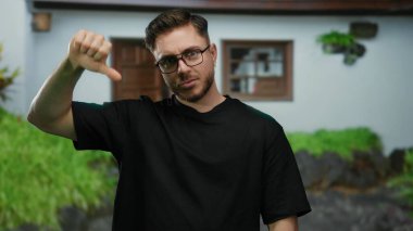 Sakallı genç bir adam şehir sokaklarında gözlüklü ve siyah tişörtlü, şehir ortamında karışık duygular ifade eden bir sokak sahnesinde başparmak kaldırıyor..