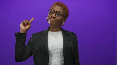 Siyah ceketli ve gözlüklü bir kadın mor stüdyoda boş bir yeri işaret ediyor..