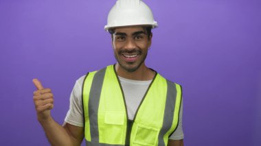 Beyaz kasketli ve sarı yansıtıcı yelek giyen İspanyol genç adam mor stüdyoda baş parmak işareti yapıyor; güven motivasyon iyimserliği başarısı.