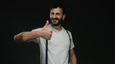 Pantolon askısı ve beyaz gömlek giyen İspanyol bir adam siyah arka plana karşı olumlu ve kendine güvenen neşeli bir gülümseme ve neşeli bir jestle başparmak kaldırıyor..
