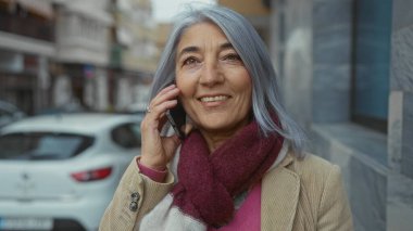 Son sınıf öğrencisi, gri saçlı bir kadın sokakta, binalarla ve park halindeki arabalarla cep telefonuyla konuşurken gülümsüyor..