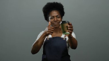 Önlük giymiş bir kadın kahve fincanı tutarken gri arka plana karşı olumlu tavır sergiliyor..