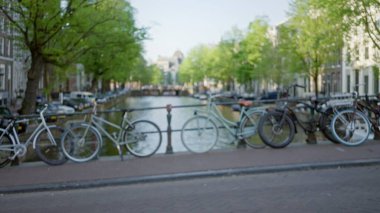 Bulanık bisikletler yeşil ağaçlar ve arka planda açık bir gökyüzünün altında tarihi mimari yapılarla dolu bir kanal boyunca dizilmiş..