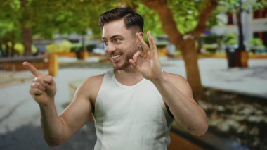 Parkta sakallı bir genç adam yeşil ağaçlar ve rahat bir atmosferle güneşli bir günde beyaz kolsuz bluz giyer ve gülümser..