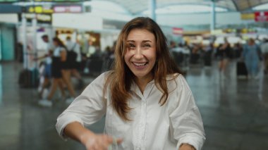 Kapalı bir havaalanı terminalinde gülümseyen genç bir kadın, arka planda canlı bir atmosfer ve çeşitli gezgin aktiviteleri sergiliyor..