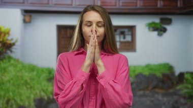 Kadın dışarıda, sakin bir bahçe ortamında meditasyon yapıyor, pembe bir gömlek giyiyor, gözleri kapalı, yeşilliklerle çevrili, sükunet ve rahatlamayı teşvik ediyor..