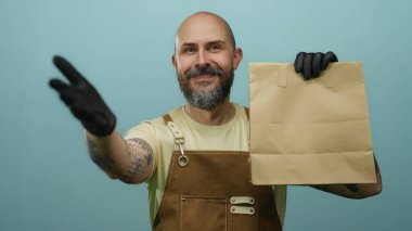 Mavi arka plan portrelerine karşı eldivenli, sakallı, kağıt çantalı kel bir adam..