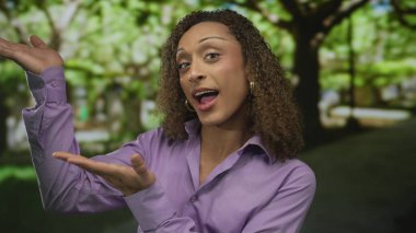 Çember küpeler ve mor gömlek giyen bulanık ağaçların arasında, açık palmiyeler arasında sunan kadın; davetiye sıcaklığı.