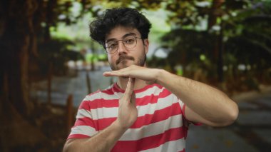 Sakallı, gözlüklü, çizgili tişörtlü, dışarıda yeşil bir parkta mola veren İspanyol bir genç..