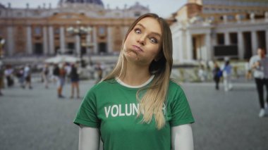 Gönüllü tişörtlü sarı saçlı kadın Aziz Peter Meydanı 'ndaki tarihi mimarisi olan Vatikan şehrinde komik surat ifadeleri sergiliyor.