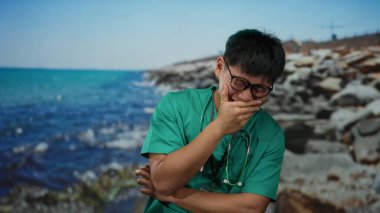 Gözlüklü ve steteskoplu genç adam kayalık bir plajda gülüyor mavi deniz arkaplanı ve berrak gökyüzü açık havada doğa ve sağlık hizmetlerini birleştiriyor..