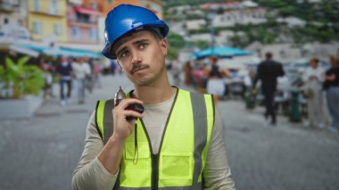 Yansıtıcı yelek ve şapka giyen genç İspanyol adam şehir caddesinde matkap tutuyor. Yoldan geçenler ve açık hava düzenlemeleri arasında kentsel inşaat işi olduğunu gösteriyor..